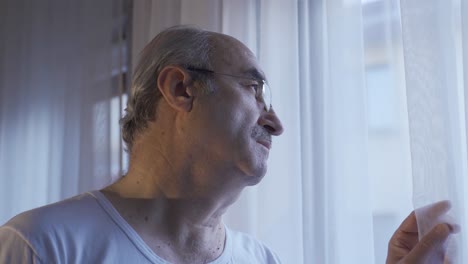 Unhappy-old-man-looking-out-the-window-in-the-living-room-at-home-takes-off-his-glasses.